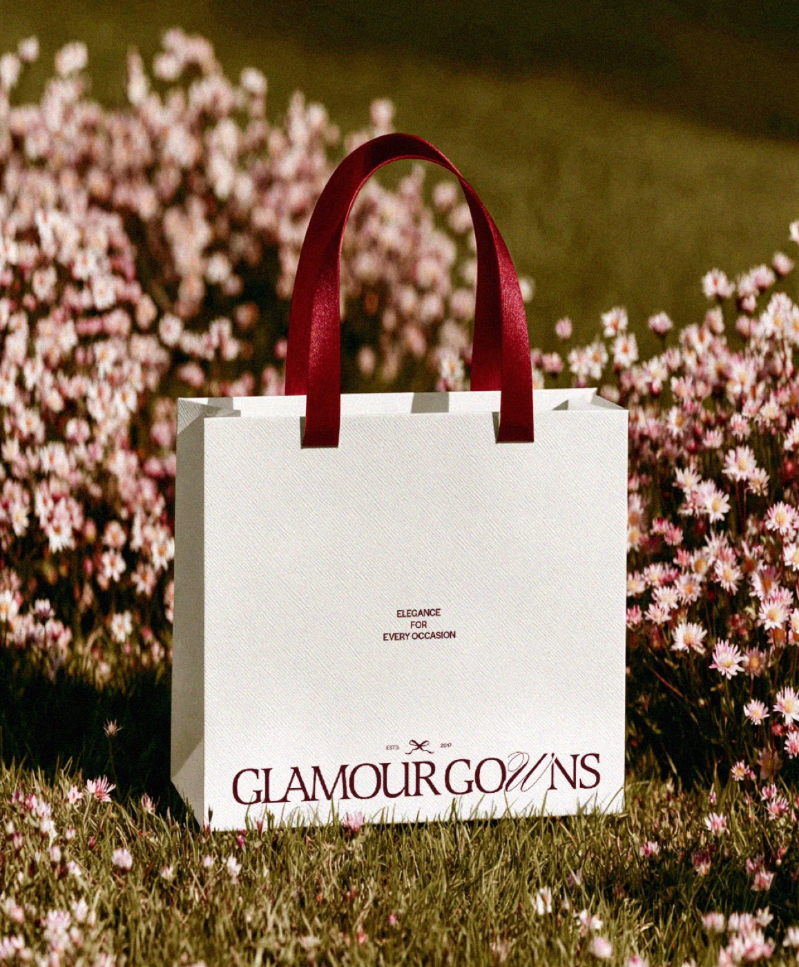 A Glamour Gowns shopping bag with burgundy handles in a field of pink flowers